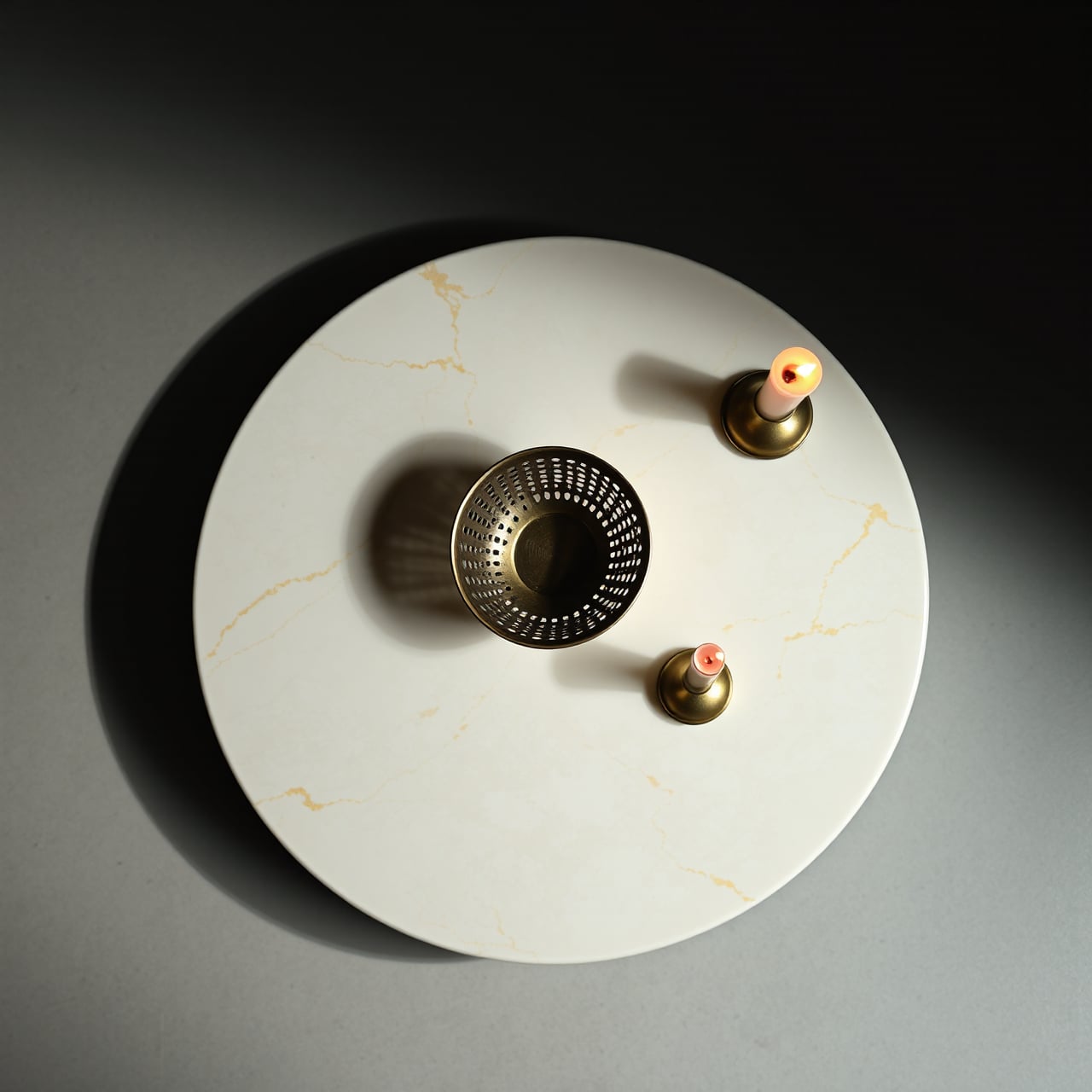 Overhead view of the round marble göbek taşı — a perforated brass bowl at center flanked by two lit brass candle holders, light raking across the gold-veined stone.