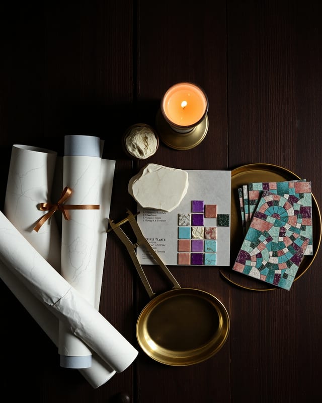 Atelier still life — rolled architectural drawings, brass calipers, marble samples, mosaic tile samples on a dark walnut table lit by a single candle