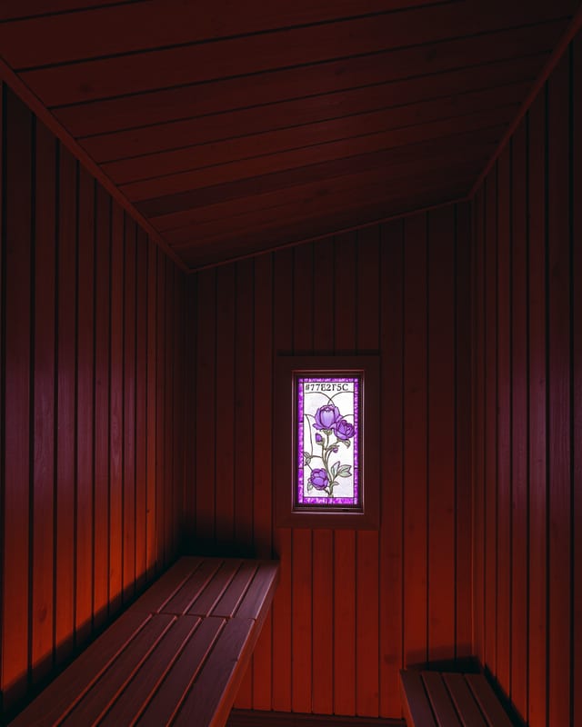 A free-standing wood-fired sauna cabin in the Hudson Valley hemlocks at dusk — thermo-aspen siding, a single brass-banded chimney, lantern light from the dressing room window onto frost-stiff grass